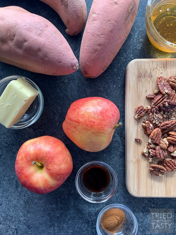 Three sweet potatoes, a bowl with butter, two gala apples, a pinch bowl of vanilla, roughly chopped pecans on a wooden cutting board & a small bowl of honey on a counter top