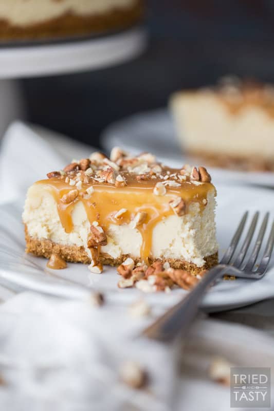 Slice of cheesecake covered in caramel & topped with pecans on a white plate with silver fork