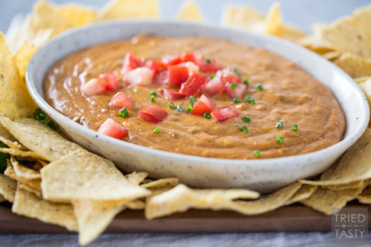 Bowl of queso in a ceramic dish surrounded by tortilla chips and topped with chopped tomato and diced chives