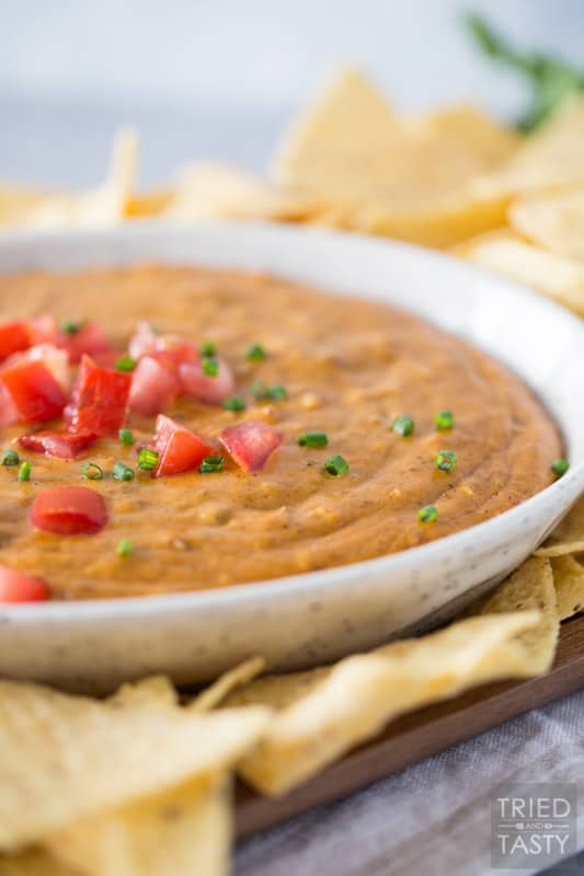 Bowl of queso in a ceramic dish surrounded by tortilla chips and topped with chopped tomato and diced chives