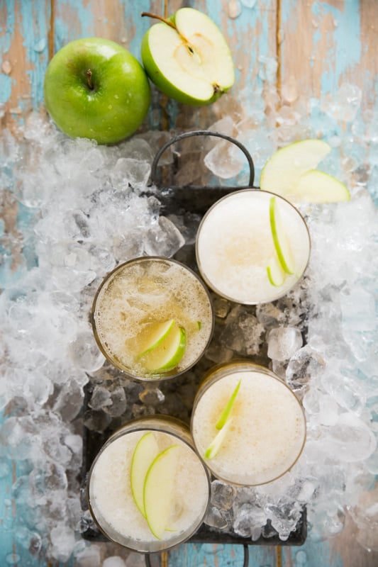 Several glasses of Green Apple Kombucha Slushy, with green apples and ice in the background.