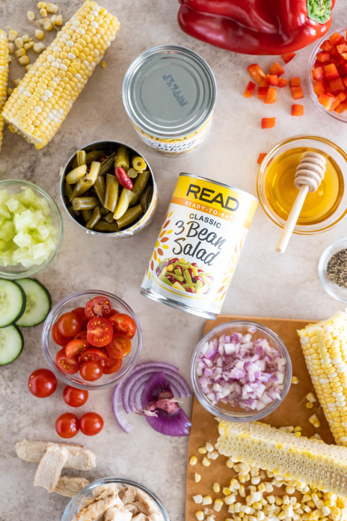 A photo of the various ingredients used in this recipe, including a can of READ 3 Bean Salad, Cherry Tomatoes, Red Onion, Corn on the Cob, Cucumber, Honey, and Chicken Breast.